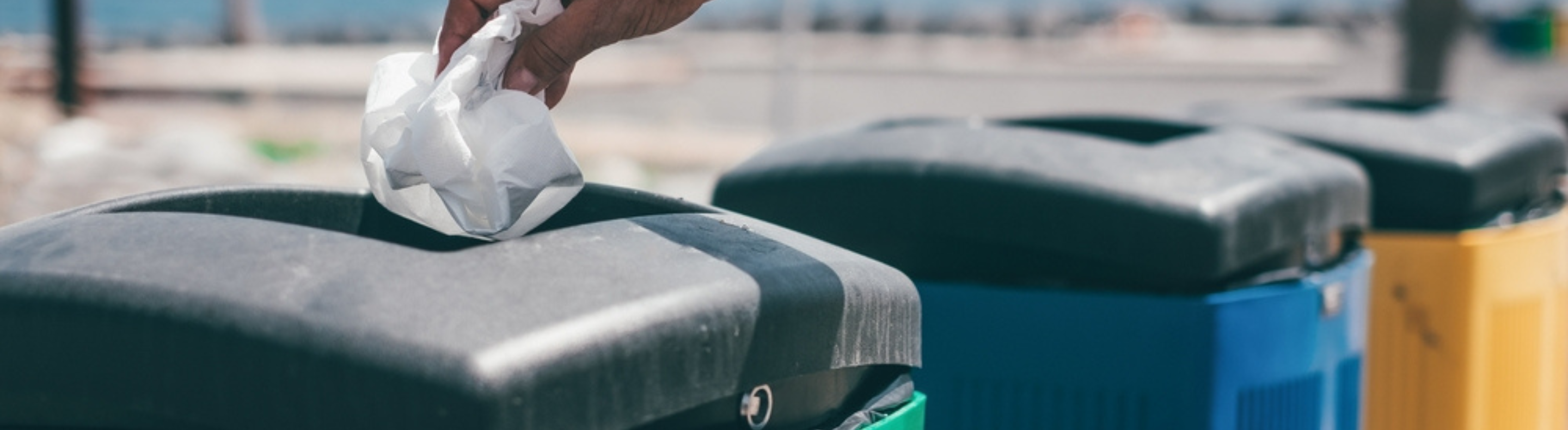 Mão descartando um resíduo em lixeira de coleta seletiva verde, com lixeiras azul e amarela ao lado.