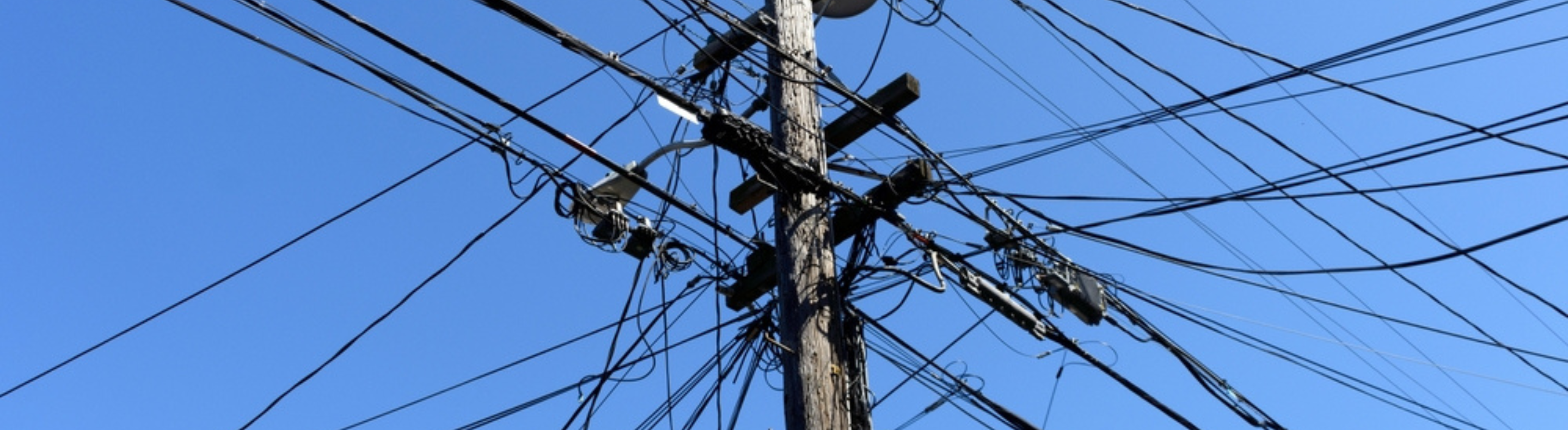 Poste de energia com grande quantidade de fios emaranhados e desorganizados. No fundo, um céu azul e sem nuvens.